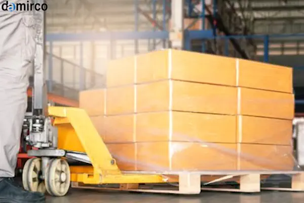 یک لیفتراک دستی نارنجی رنگ در حال جابجایی یک پالت چوبی در یک انبار صنعتی